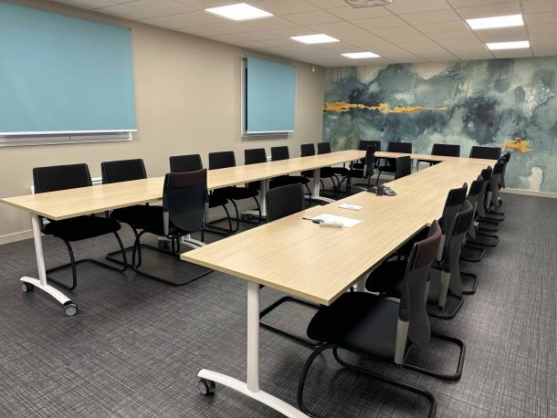 Salle de réunion spacieuse équipée de longues tables modulables en bois clair, entourées de chaises noires, avec un mur décoratif à motifs aquatiques bleus et dorés, et des stores bleus sur les fenêtres.