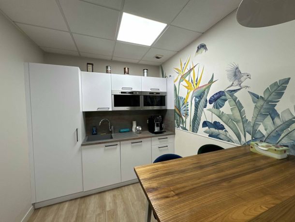Salle de pause aménagée avec kitchenette blanche sur mesure, table en bois, et un mur décoré d’une fresque tropicale avec des motifs de feuillages et d’oiseaux, offrant un espace convivial et moderne.